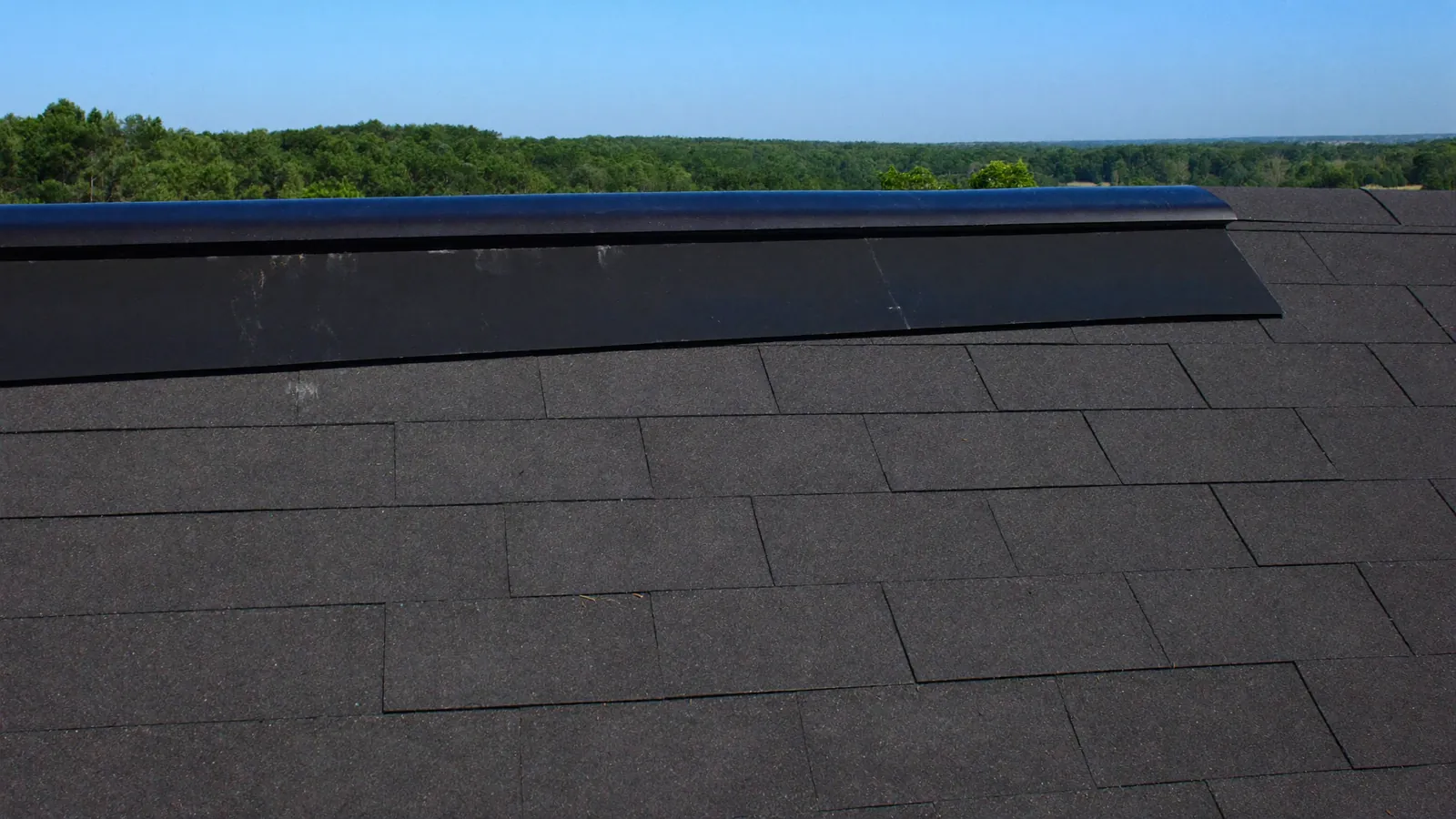 Continuous ridge vent running along the peak of a shingle roof, with the ridge cap shingles installed over it against a blue sky