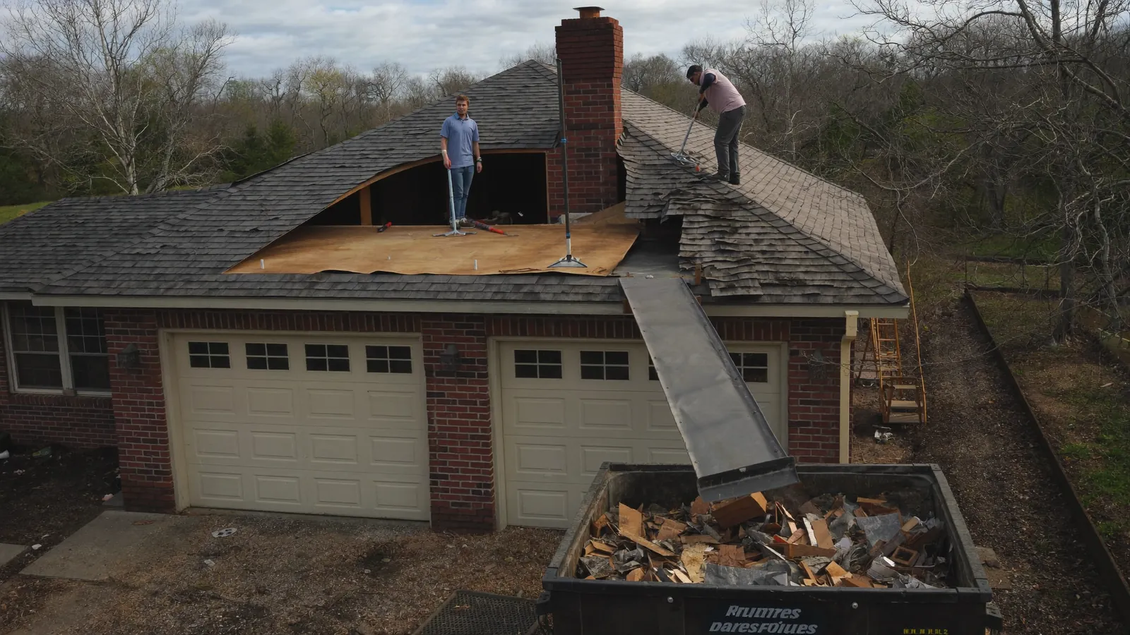 Crew tearing old three-tab shingles off a roof deck down to the bare plywood, with a dump trailer parked below on the driveway of a Tennessee home